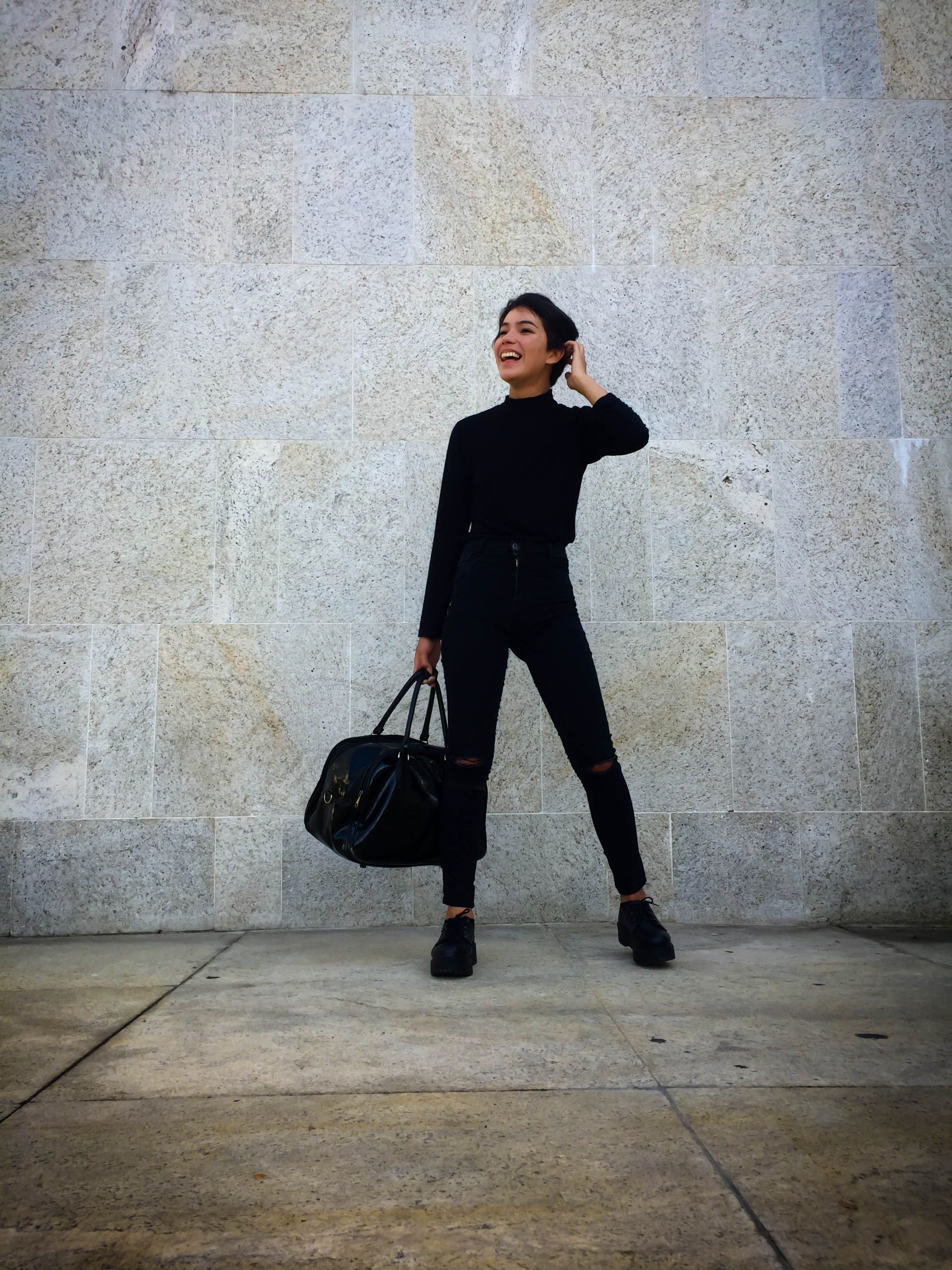 Full-length portrait of a fashionable woman in black standing against a stone wall, exuding confidence and style.