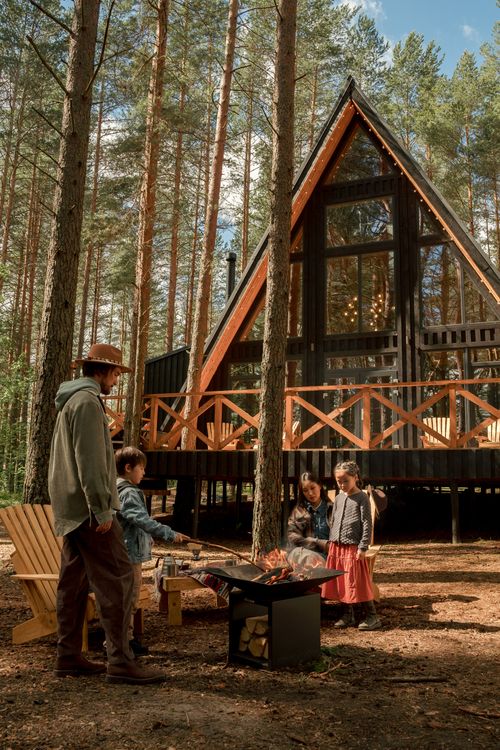 Free A family gathering around a campfire outside a rustic A-frame cabin in a forest setting, creating warmth and bonding. Stock Photo
