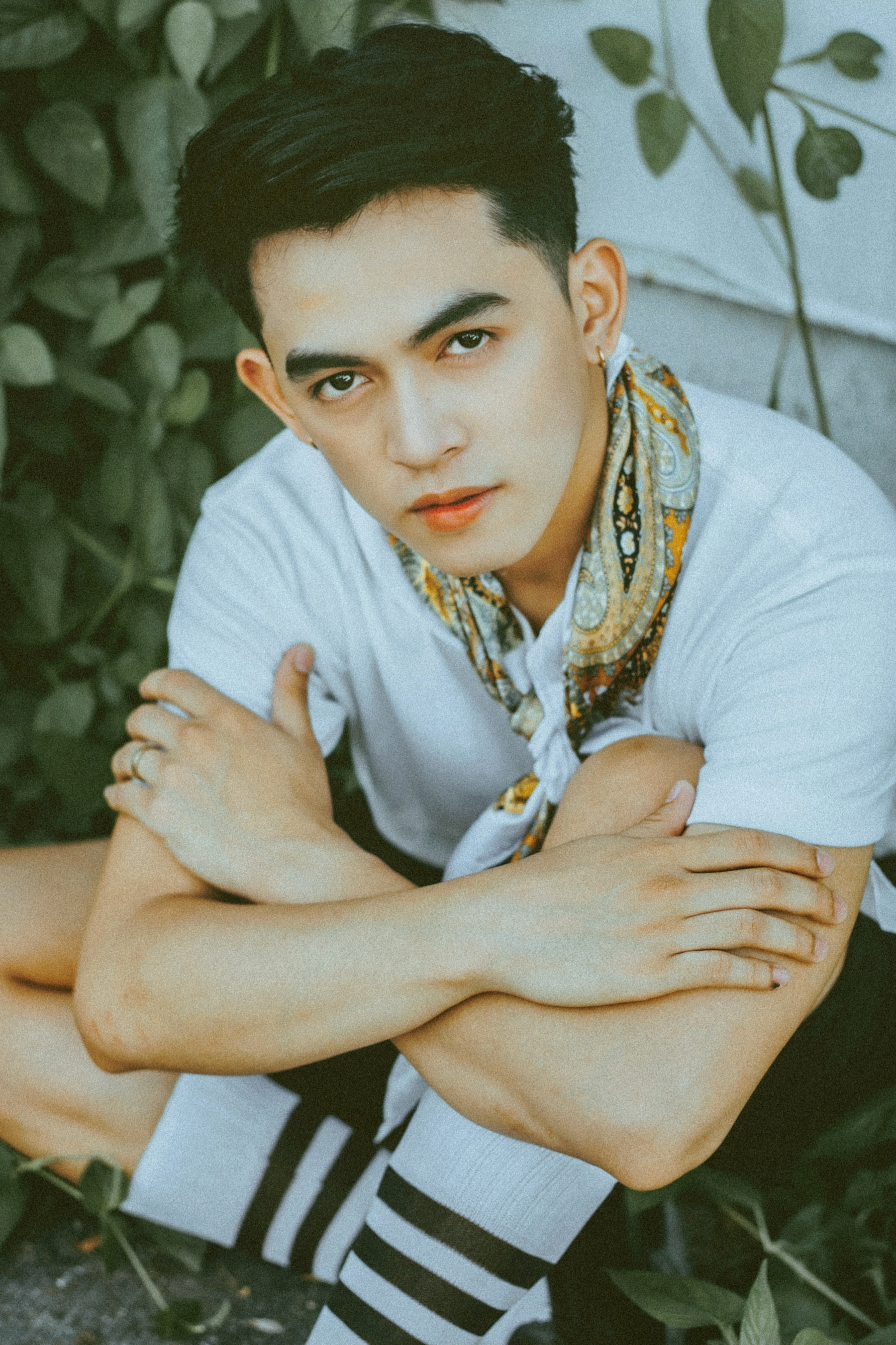 Serious young man with a scarf and rings posing thoughtfully outdoors among greenery.