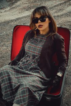 Fashionable woman sitting in open suitcase smoking, wearing sunglasses in outdoor setting.