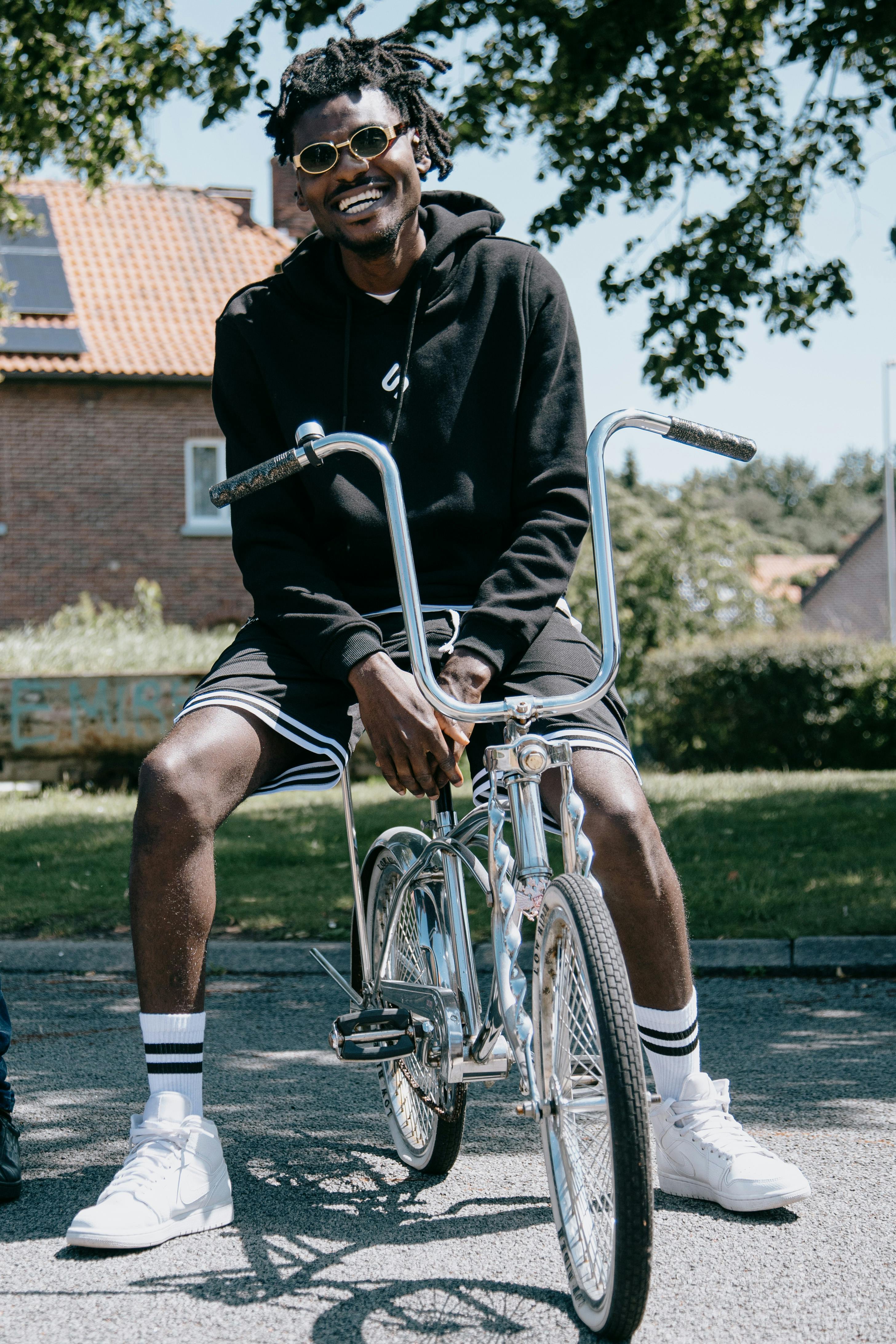 African American man sitting on a bike outside, smiling in casual attire under sunny skies.