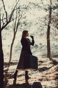 A stylish woman carrying a suitcase walks through a sunlit autumn forest.