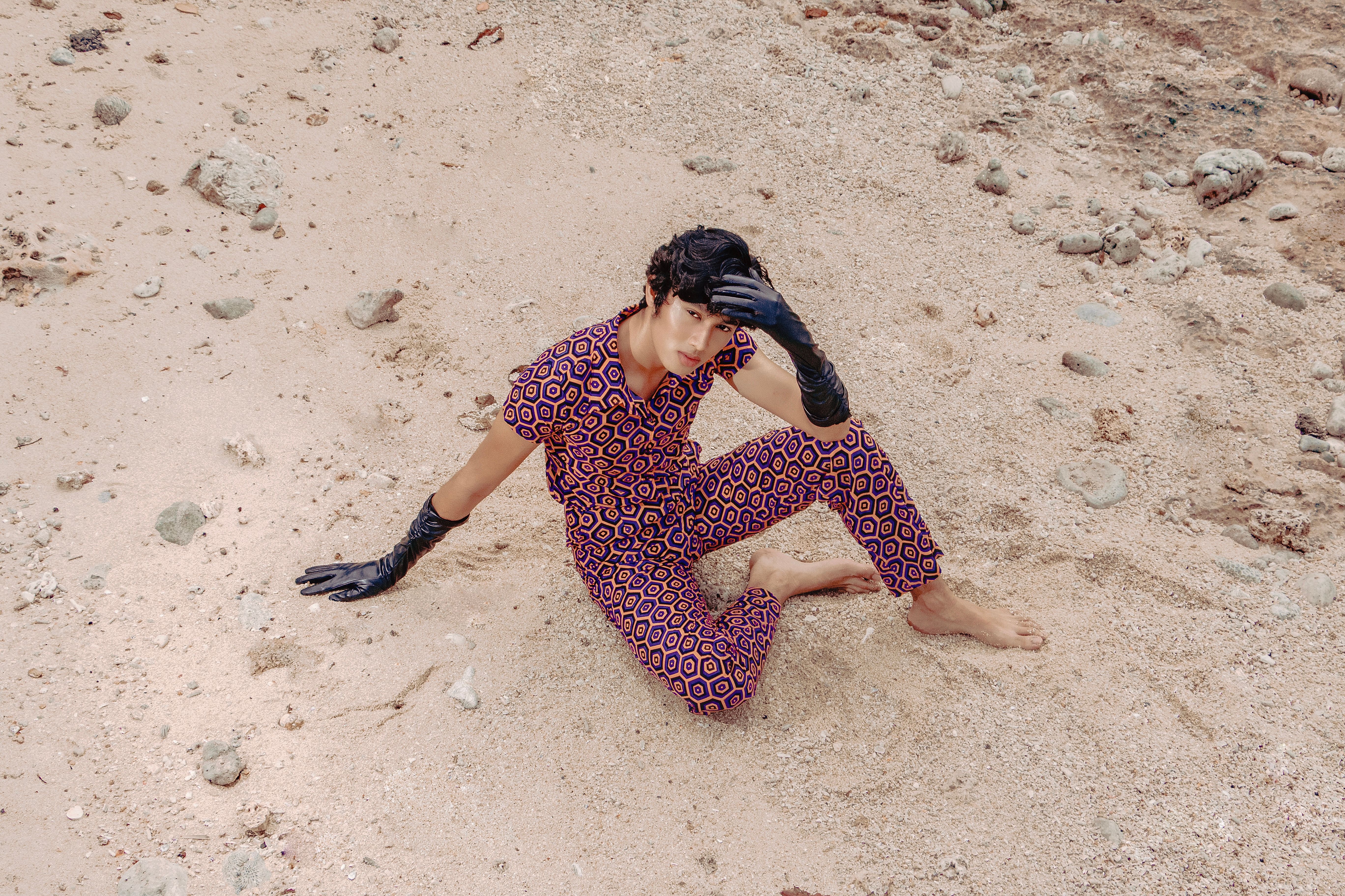 Stylish Asian man in patterned outfit sitting on a sandy beach, high angle view.