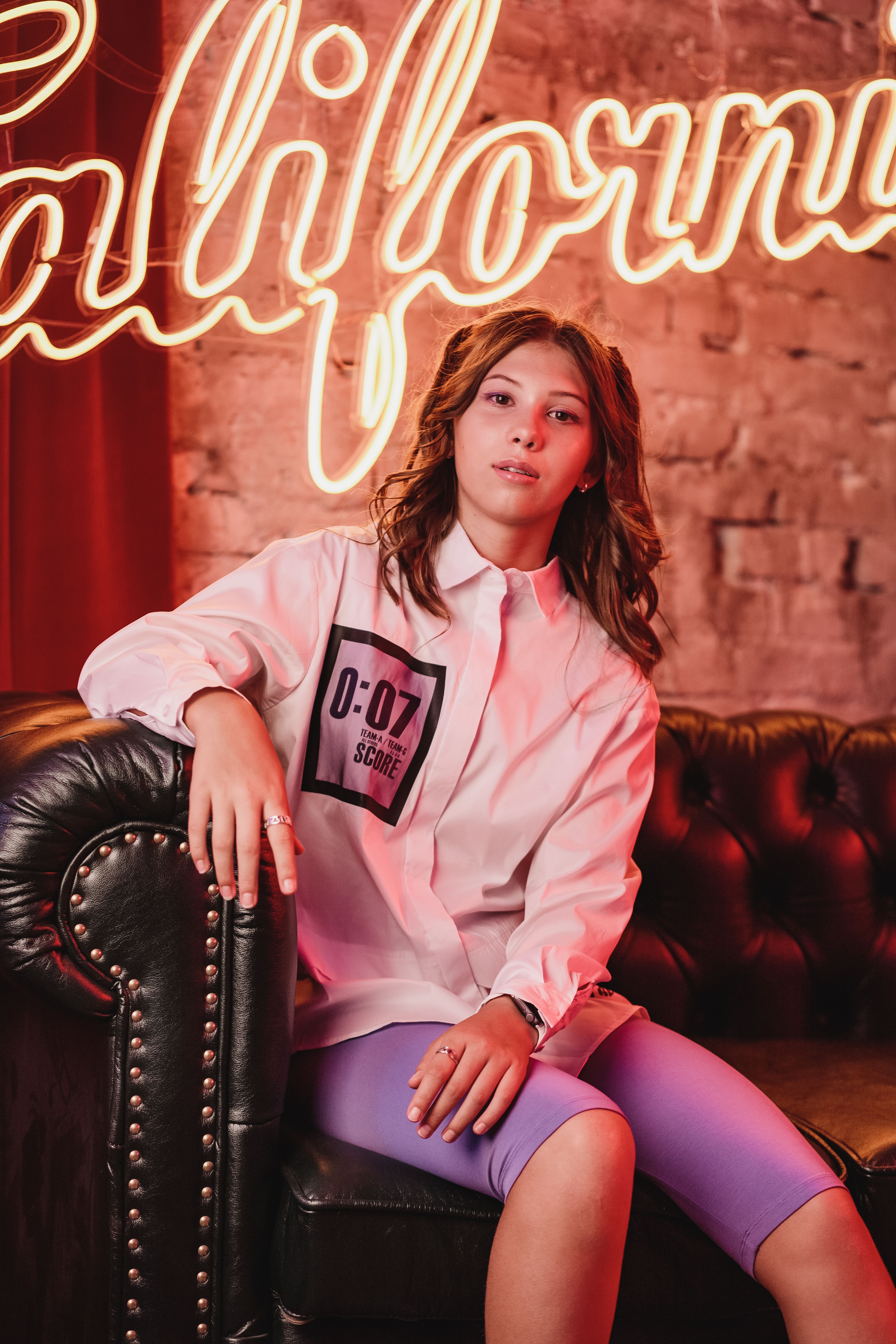 Confident young woman sitting on a leather sofa with neon California sign behind her.