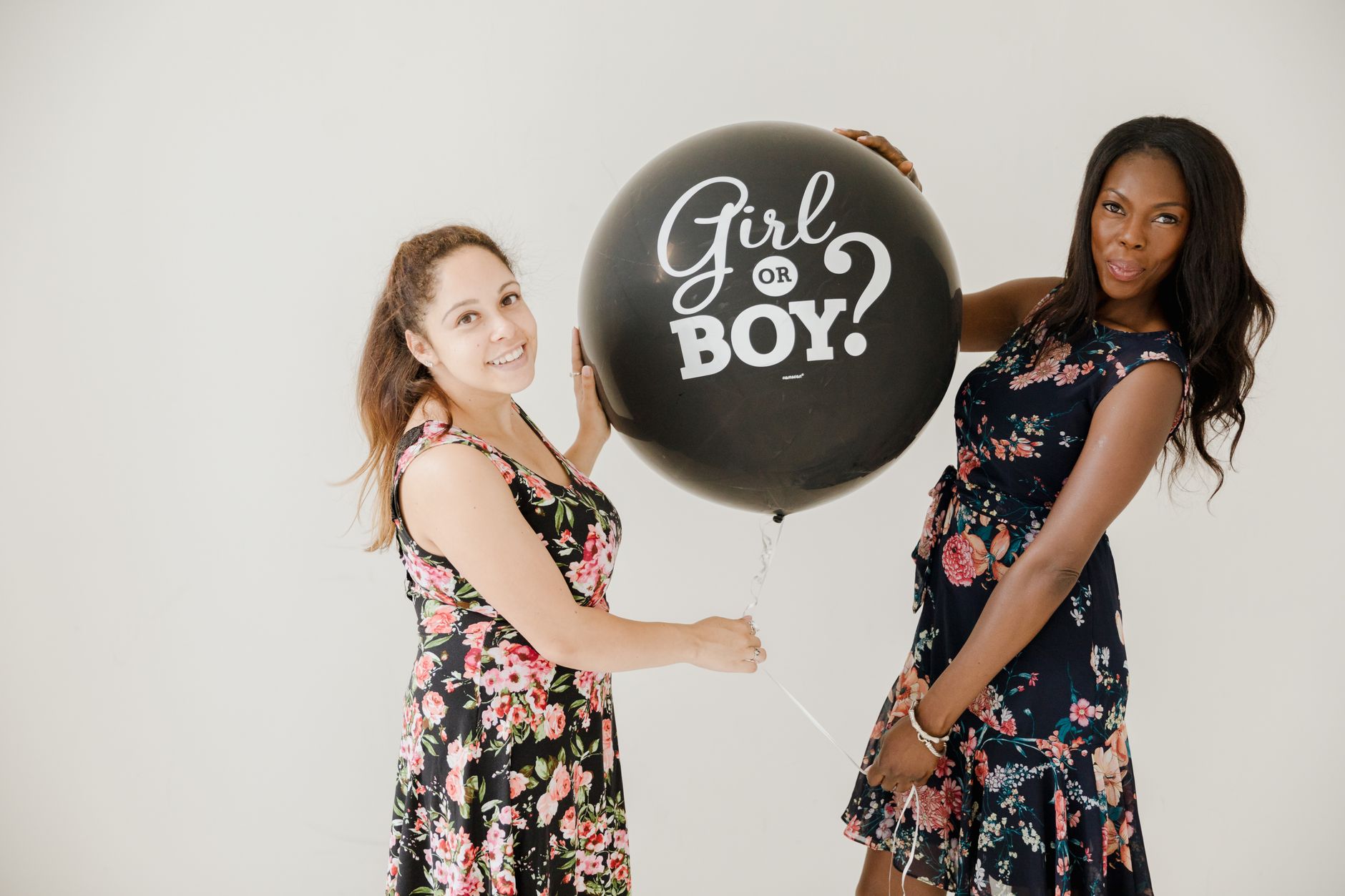Two women holding a black gender reveal balloon, wearing floral dresses on a white background.