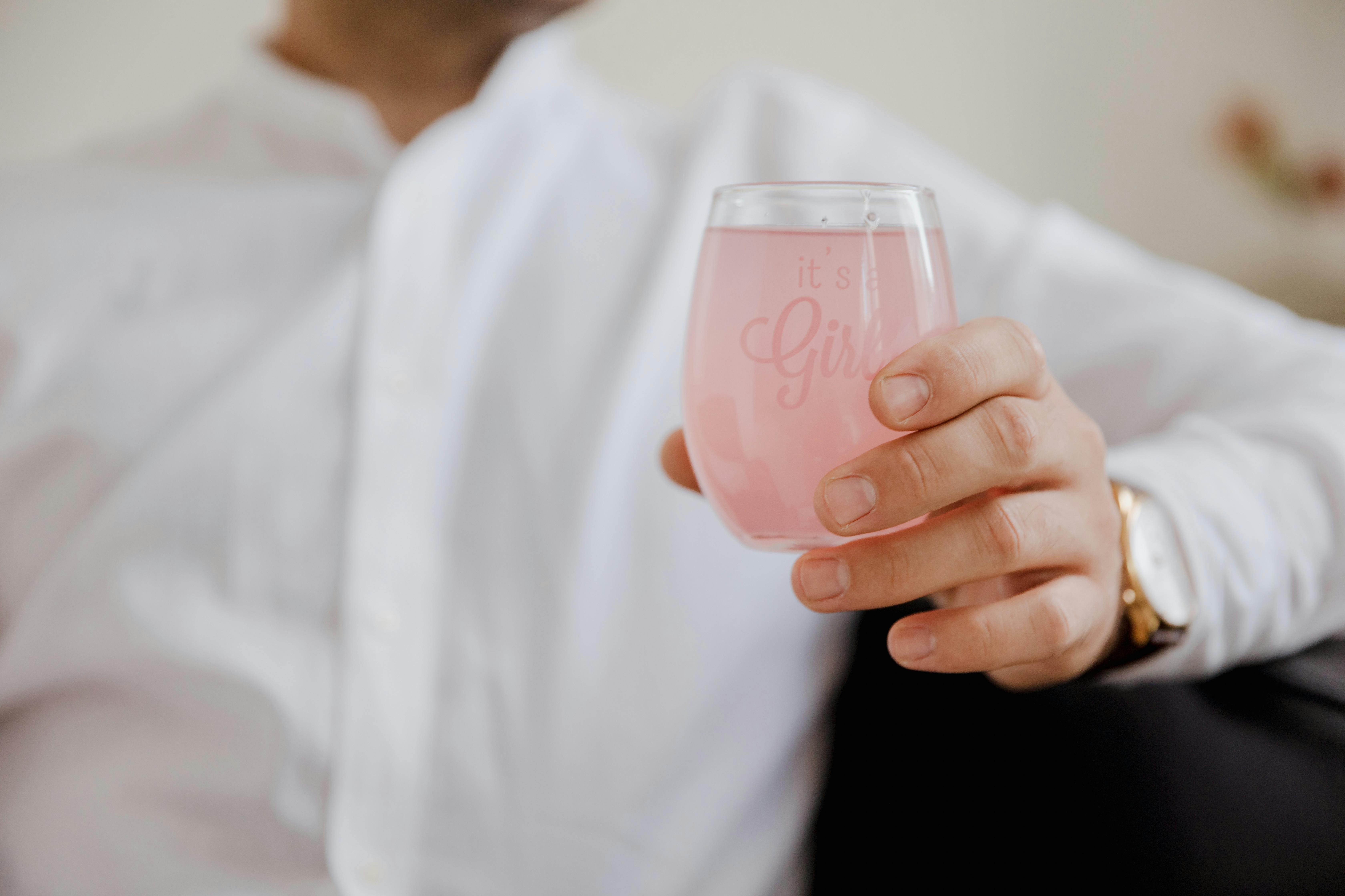 A close-up of a man holding a pink drink in a glass with 'it's a girl' text, symbolizing a baby girl announcement.