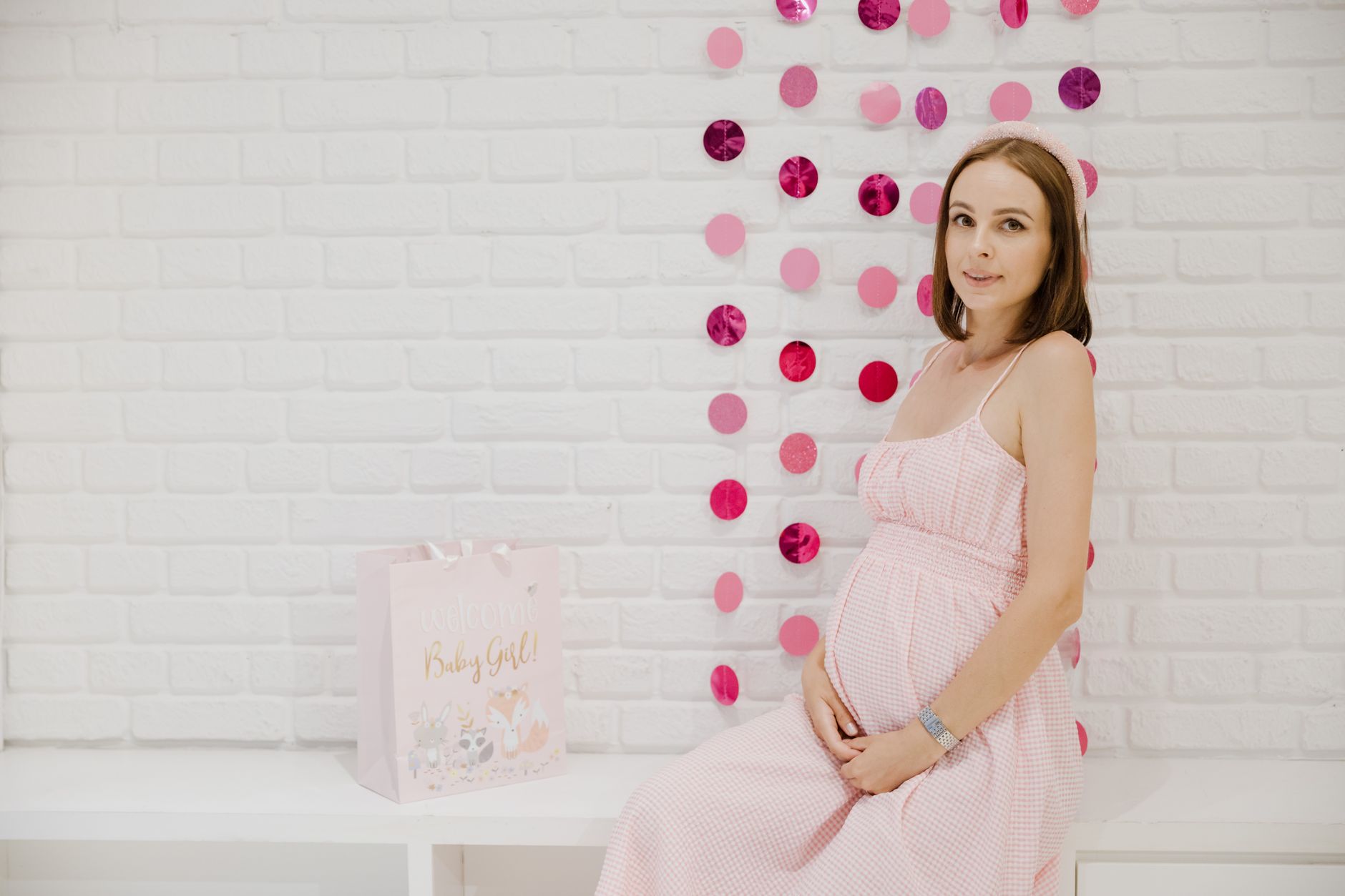 Pregnant woman in pink dress sitting by decor during a baby shower with gift bag.