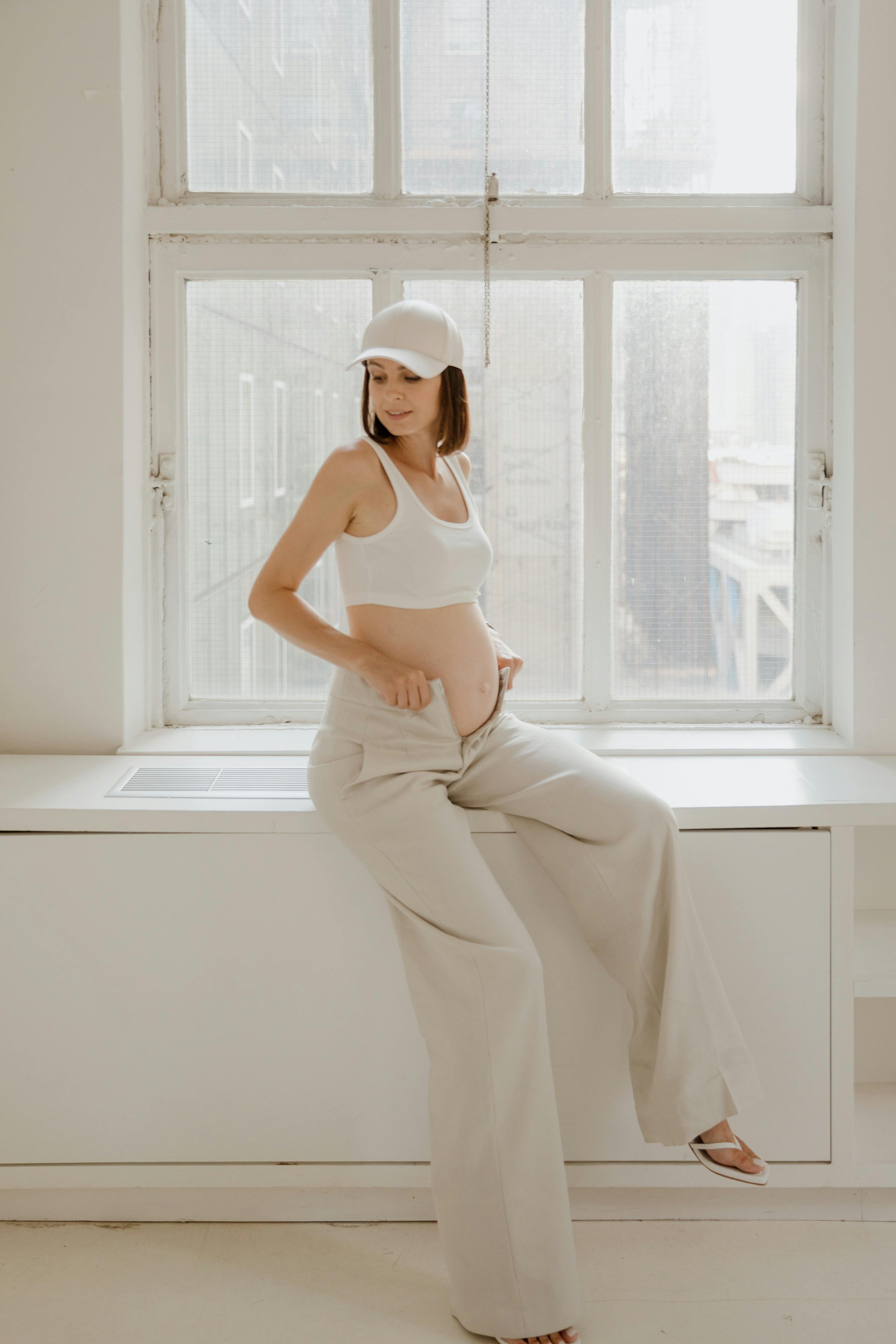 A pregnant woman in casual attire sits peacefully by an indoor window.
