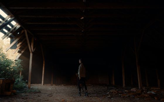 A woman stands in a dimly lit, abandoned barn with light streaming through gaps.
