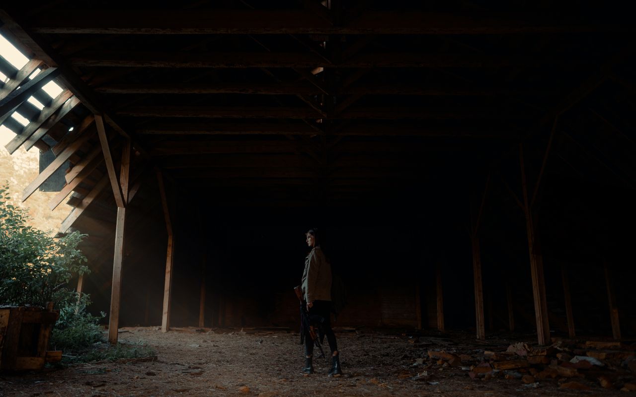 a woman stands in a dimly lit abandoned barn with light streaming thro