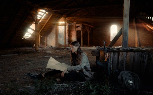 Woman Reading a Map Sitting in the Attic of an Abandoned Building with a Rifle and a Backpack