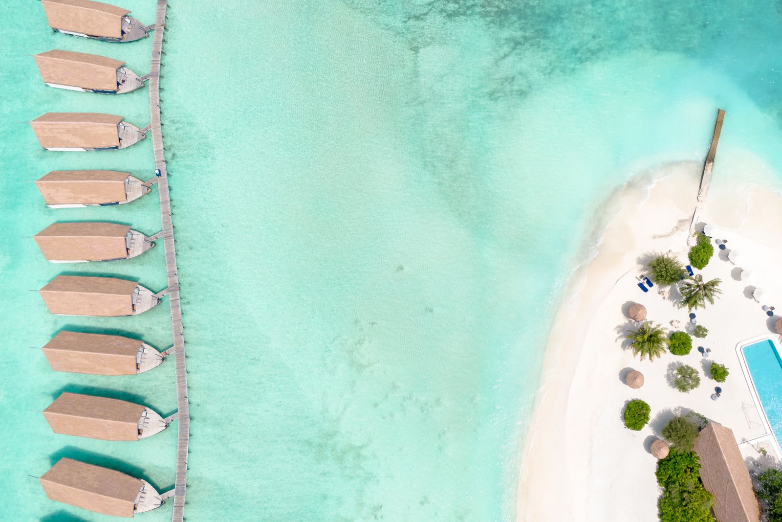 A row of overwater villas on a jetty above a turquoise lagoon beside a white-sand beach