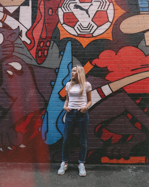 Free Woman in White Shirt and Blue Denim Jeans Standing Beside Red Wall Stock Photo