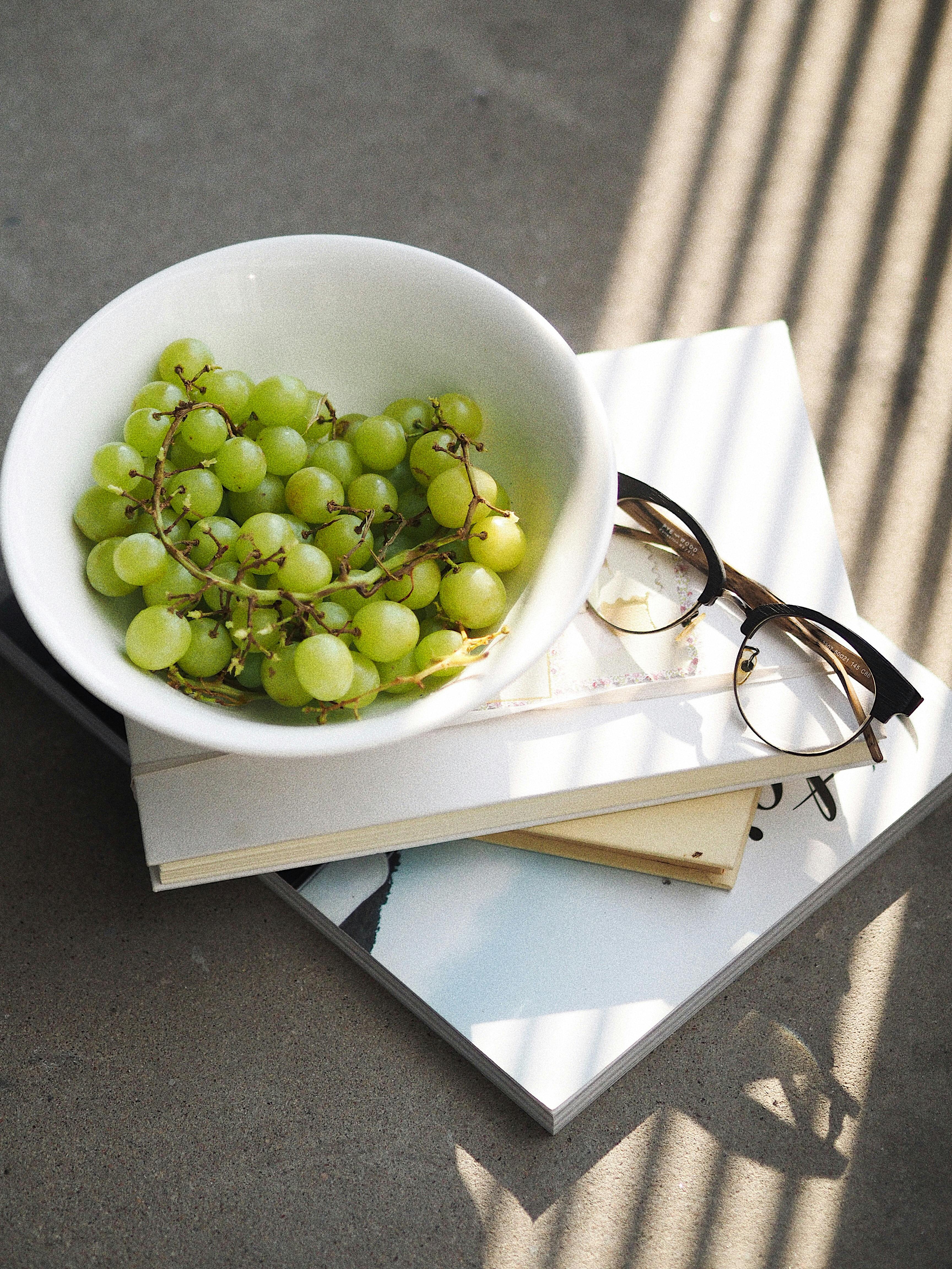 Fresh green grapes in a bowl on books with eyeglasses, under natural daylight.