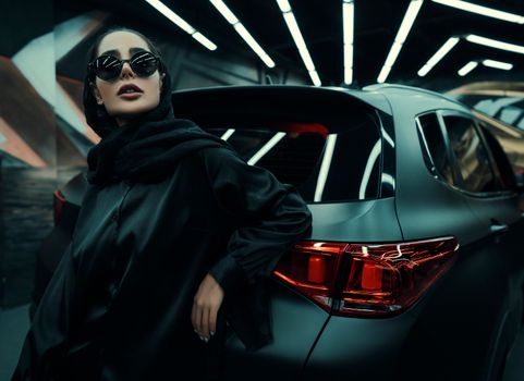 Elegant woman in sunglasses leaning against a luxury car in a modern garage setting.