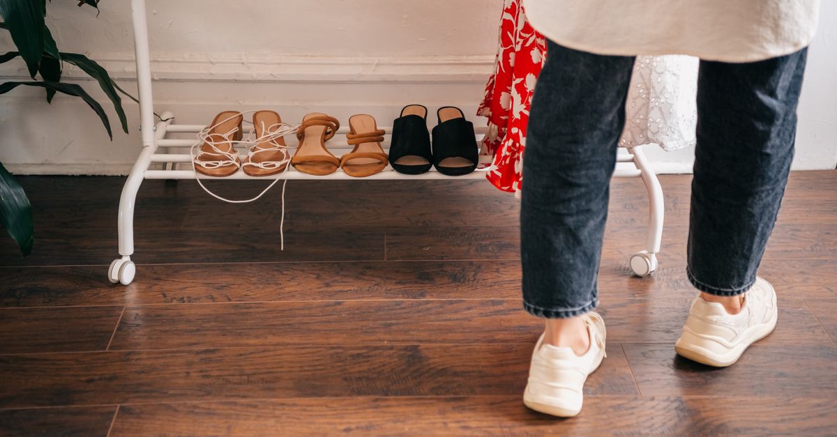 Person in casual attire browsing shoes on a rack indoors, wooden flooring visible.