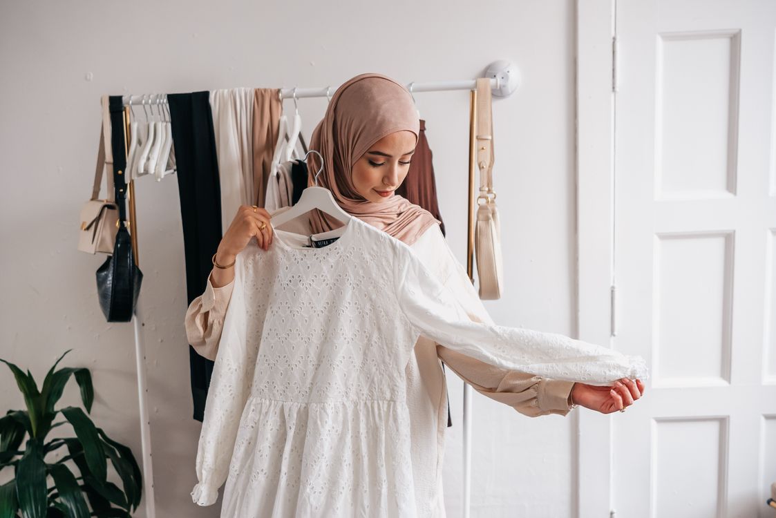 Free A Muslim woman in a hijab examines a white dress in a clothing boutique with racks of clothes. Stock Photo