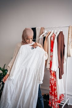 Muslim woman in hijab selects a white dress from a clothing rack indoors.