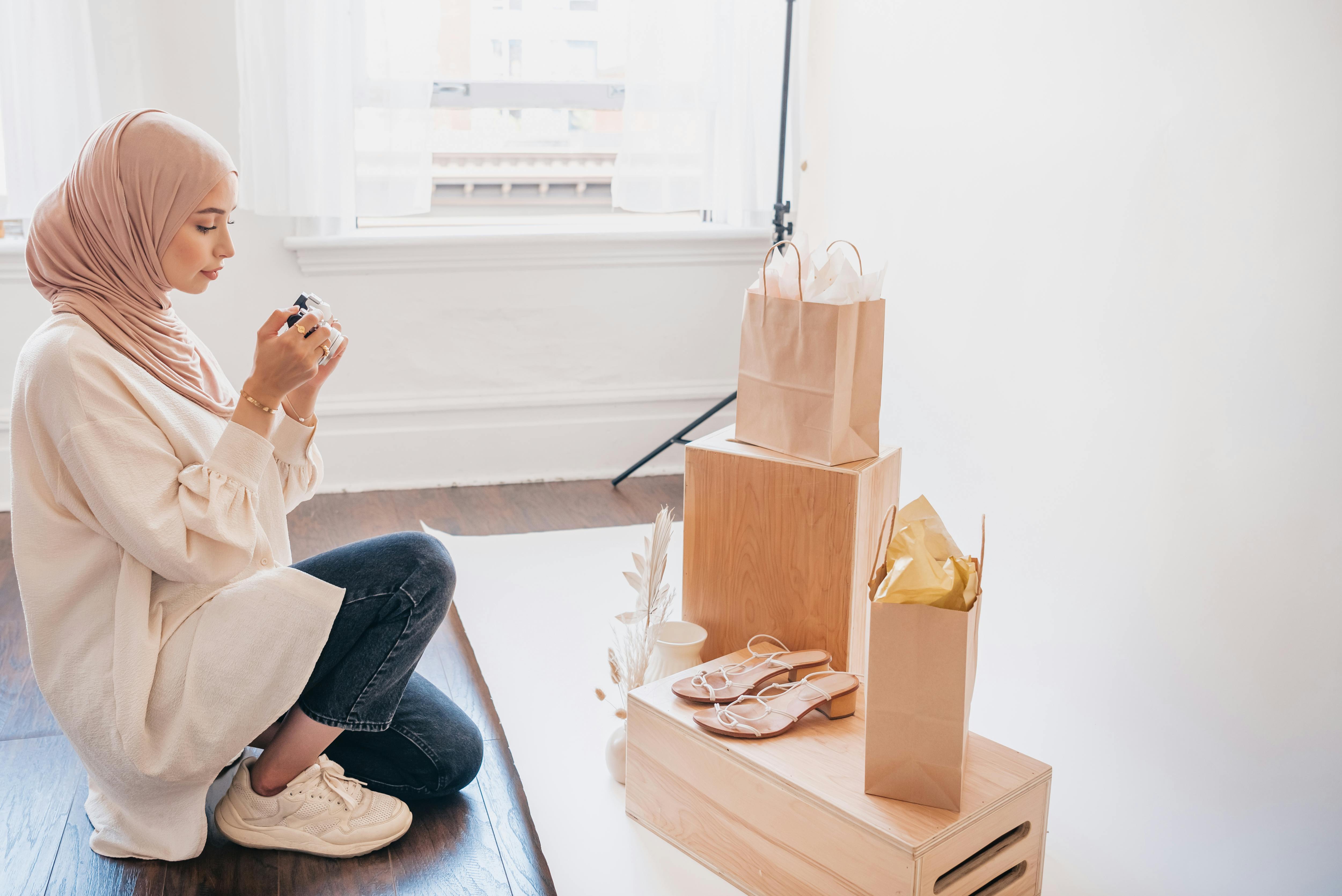 A woman in a hijab kneels to photograph a shopping display with bags and shoes.