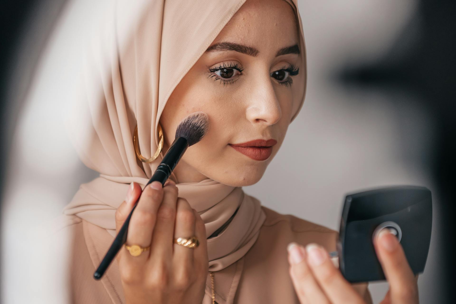 A Middle Eastern woman in a hijab applying makeup using a brush and compact mirror.