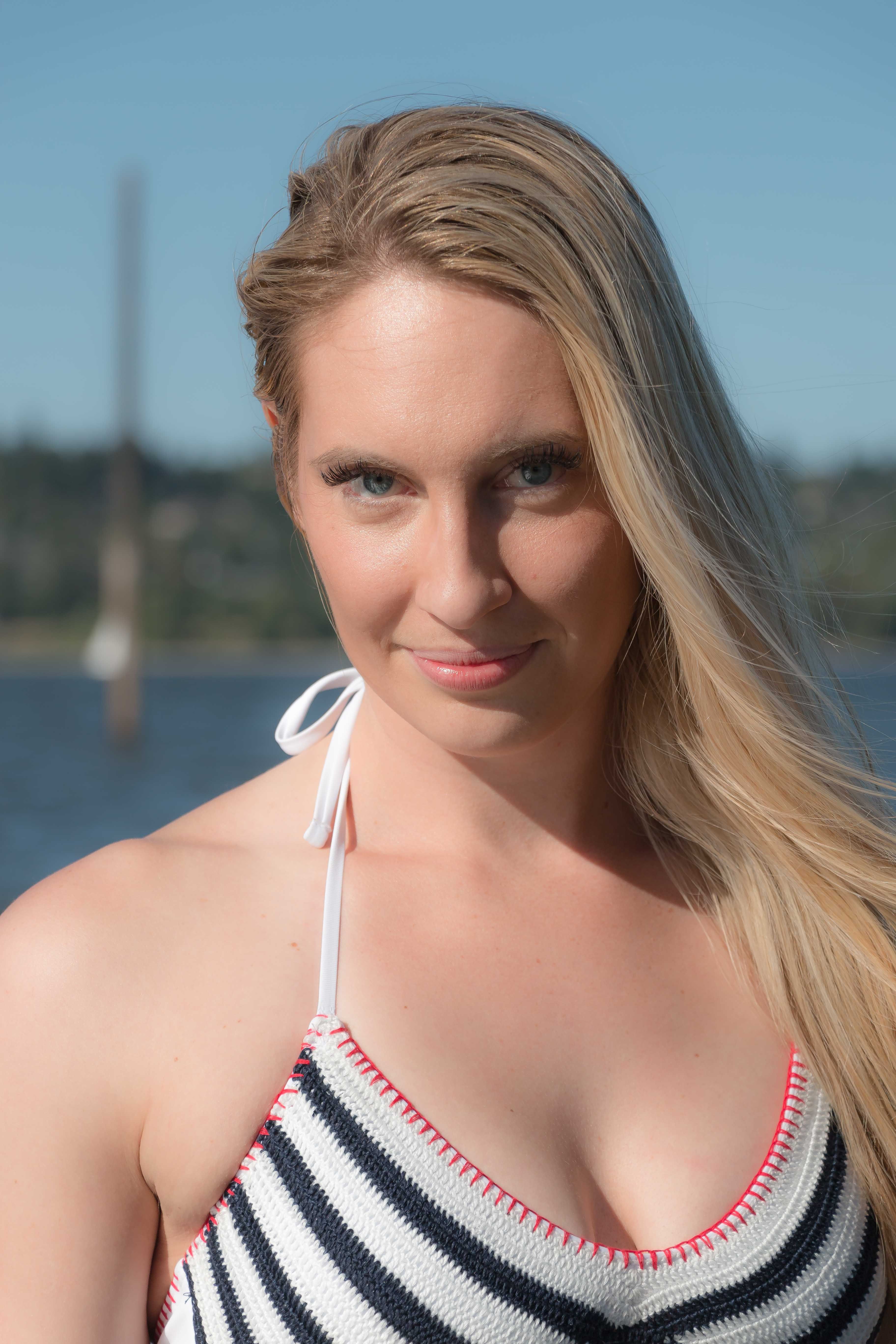 Blonde woman in summer attire posing by the water, exuding confidence.