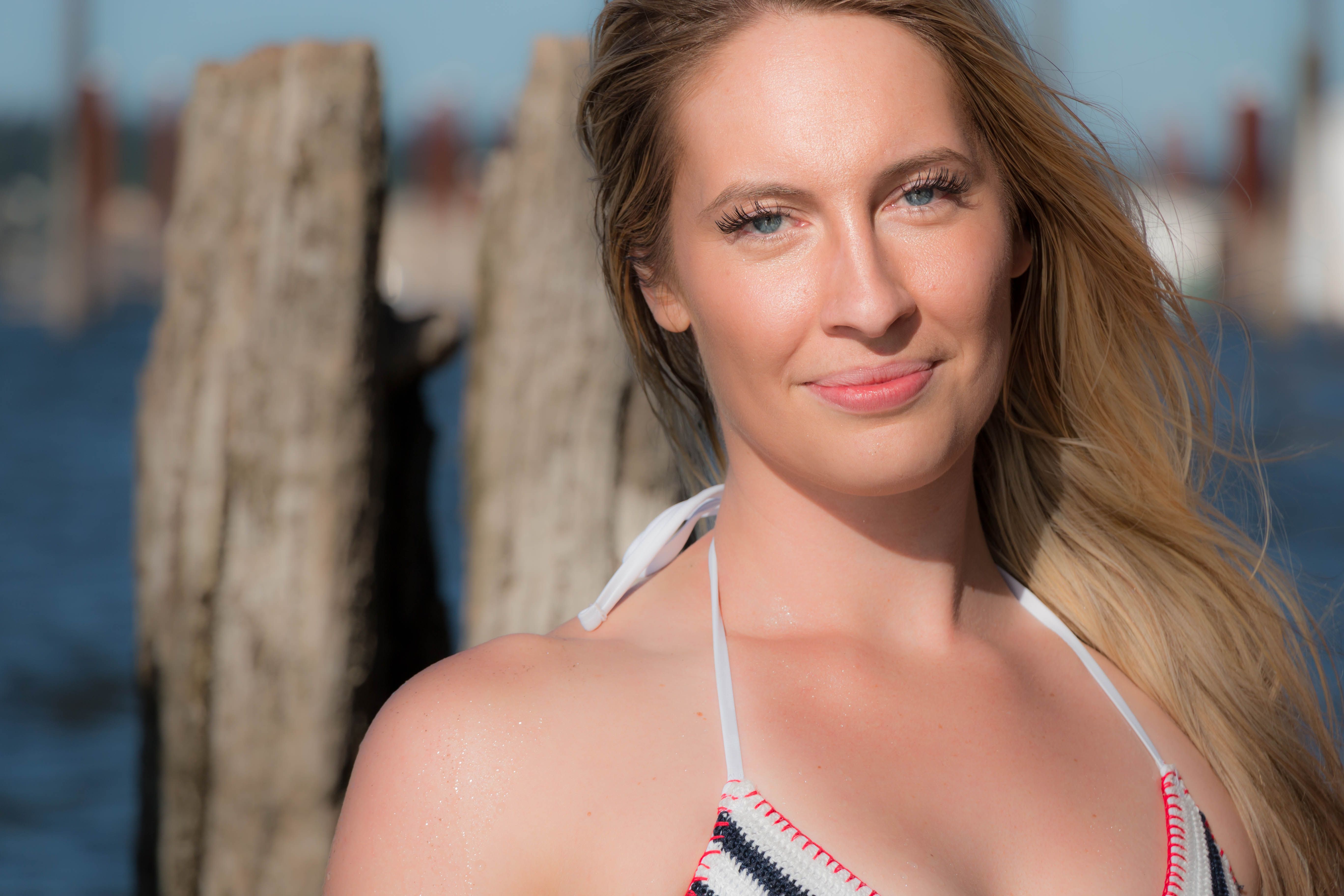 A smiling woman in summer attire posing by the waterfront under bright sunlight.