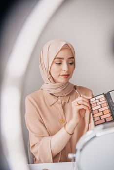 Woman in hijab applying makeup using an eyeshadow palette and mirror.