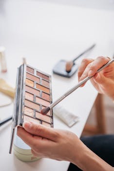 A close-up of hands holding an eyeshadow palette with a brush indoors.