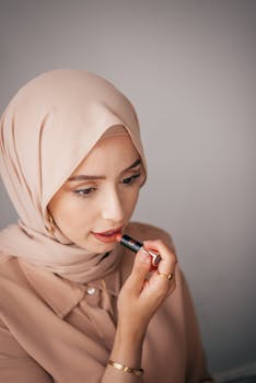 A stylish woman in a hijab applies lipstick, showcasing beauty and fashion.