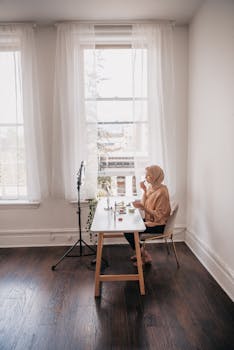 A woman in a hijab sits at a desk by the window in a room, working on her laptop in natural light.