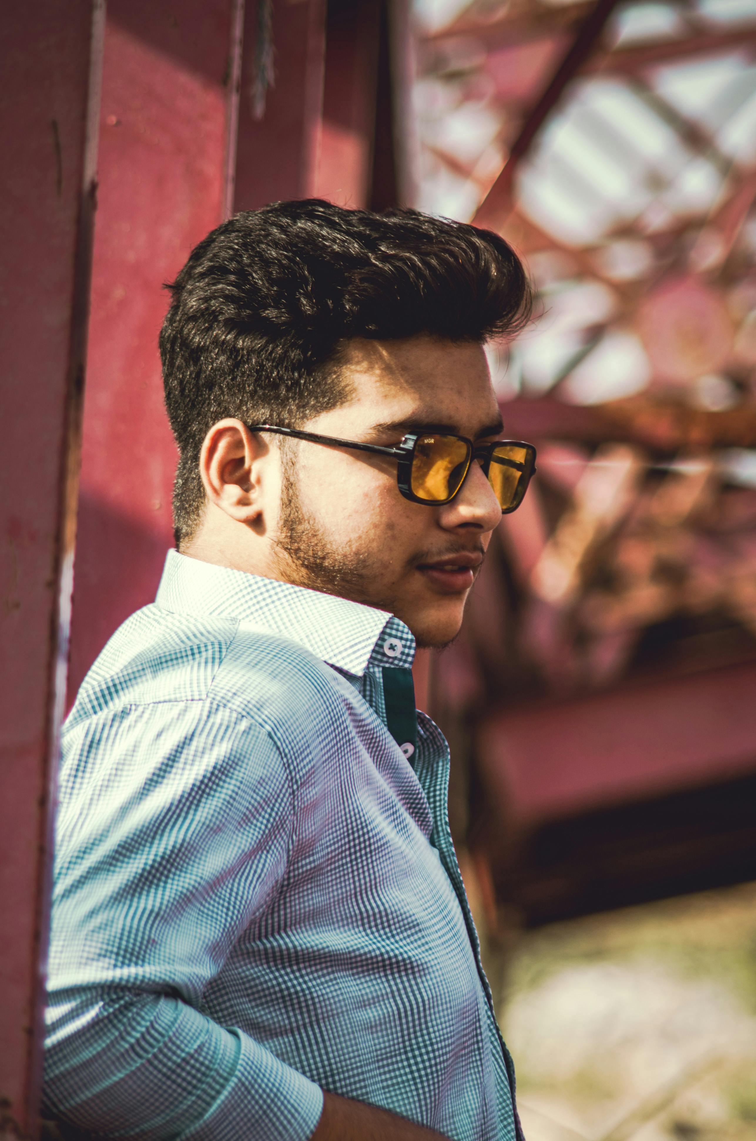 Profile of a stylish man in sunglasses leaning by a rustic structure, under a sunny sky.