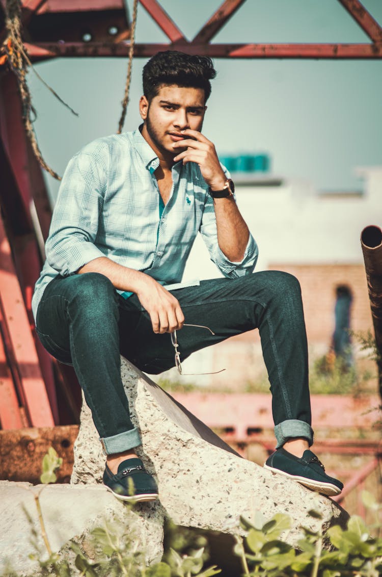 A Man In Blue Dress Shirt And Denim Pants Sitting On Rock