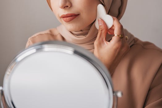 Close-up of a woman in a hijab applying makeup using a facial sponge in front of a mirror.