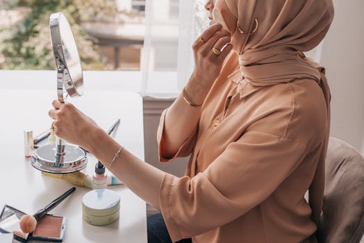 A woman in a hijab applies makeup at a bright indoor setting by a window.