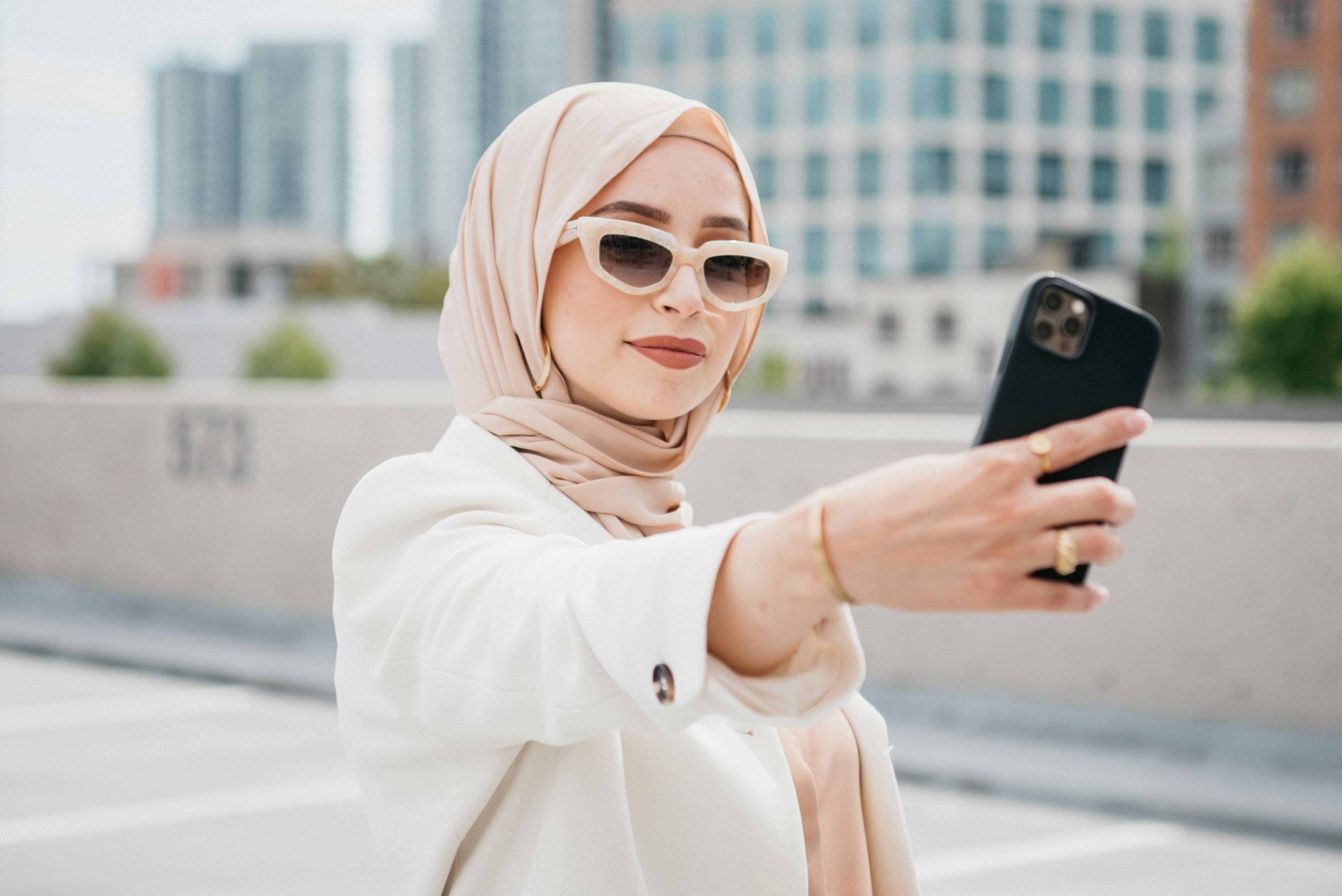 Stylish woman in hijab with sunglasses taking a selfie in an urban setting.