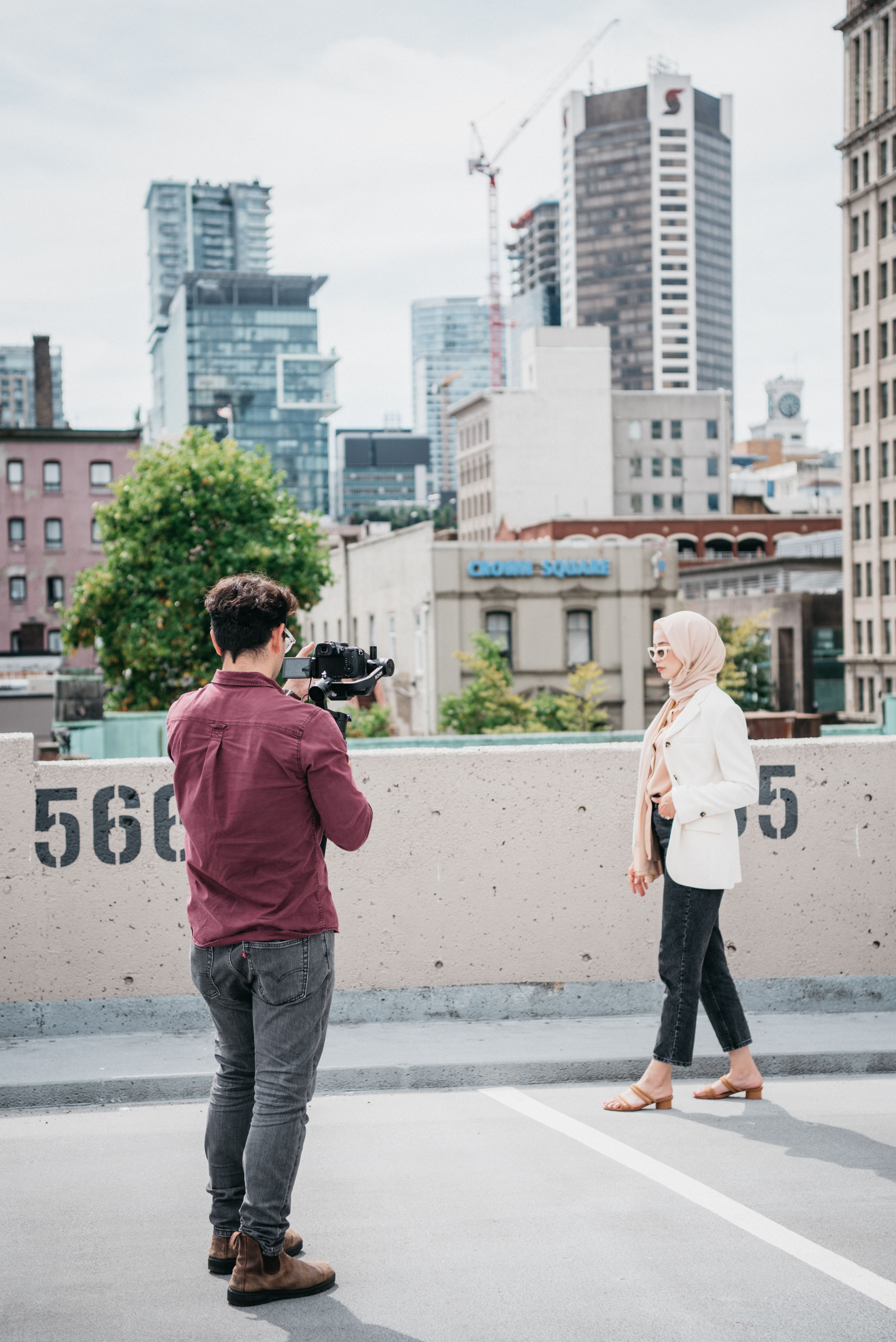 Man photographing woman in hijab on urban rooftop with city background.