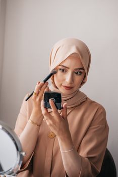 Young woman with headscarf applying makeup indoors, creating an elegant look through careful artistry.