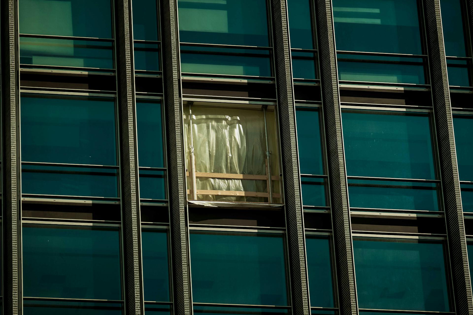 A close-up view of a modern skyscraper's facade with a focus on a broken window covered with a temporary plastic sheet.