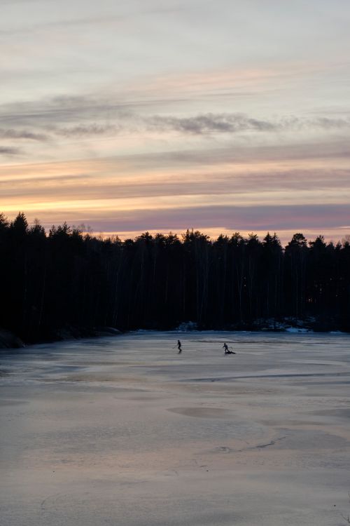 Free Scenic frozen lake at sunset with skaters silhouetted against colorful winter sky. Stock Photo