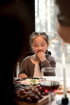 A young girl eats at a cozy, candlelit dinner table with a relaxed and joyful expression.