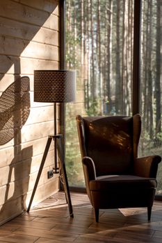 Sunlit armchair and lamp in a wooden cabin interior, overlooking a serene forest view through a glass window.