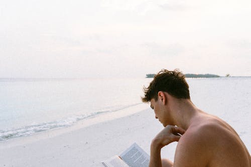 Free Topless Man Reading Book Beim Sitzen Am Strand Stock Photo