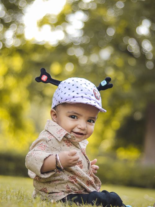 Free Adorable baby boy sitting on grass, wearing a patterned hat with playful design, in a sunny park. Stock Photo