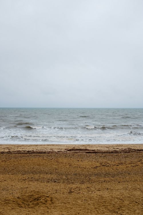 Free A serene beach scene with overcast skies and gentle waves on a sandy shore. Stock Photo