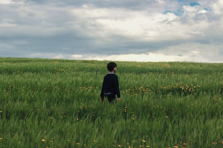 Un niño en un suéter negro camina por un campo verde y exuberante bajo un cielo nublado.