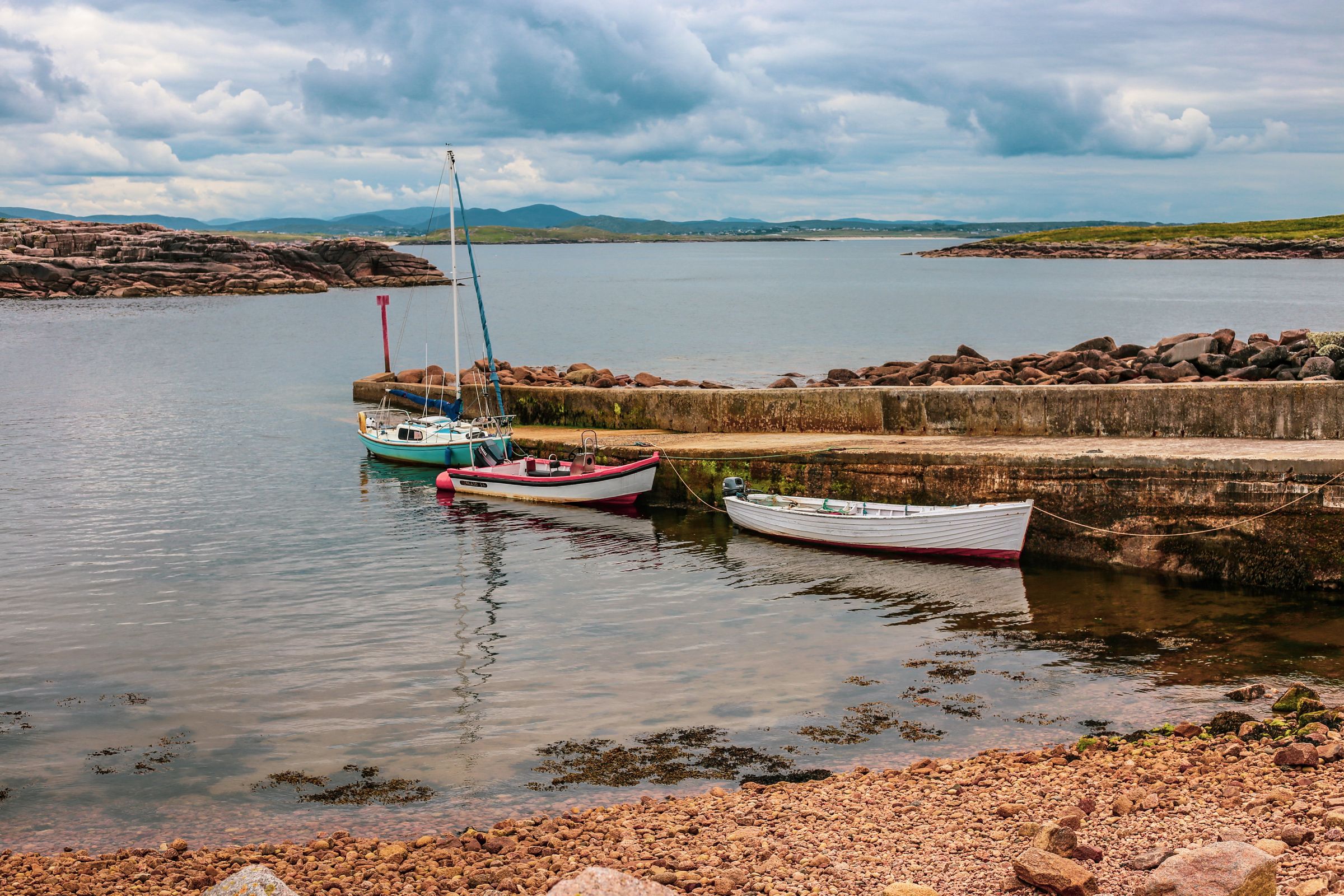 A peaceful harbor scene in Ireland featuring three moored boats against a rocky shoreline under a cloudy sky
