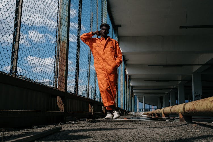 A Low Angle Shot Of A Man In Orange Jumpsuit Standing Near The Chain Link Fence