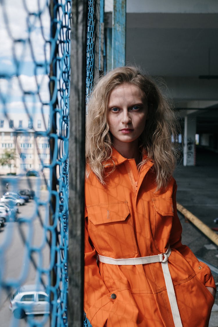 Woman In Orange Jumpsuit Leaning On Wire Mesh Fence