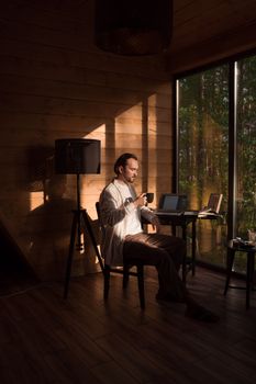 Man enjoying a coffee break while working from a cozy modern cabin with sunlight streaming through the windows.