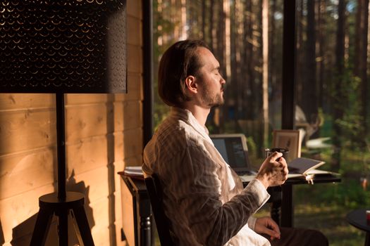 A man enjoying coffee while working remotely in a sunlit cabin, surrounded by nature.